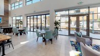 Located at 2205 Natomas Park Drive in Sacramento, CA, this single-story community clubhouse delivers a polished, contemporary environment designed for both social gatherings and everyday use. The space is defined by its soaring double-height ceilings, floor-to-ceiling glass walls, and large-format light tile flooring that amplifies the abundance of natural light throughout.

The main gathering area features a spacious lounge and dining zone furnished with tufted seating, mint-green dining chairs, and colorful accent rugs that add vibrancy to the neutral palette. Multiple sets of full-height sliding and hinged glass doors open directly onto a well-appointed outdoor courtyard, creating a seamless indoor-outdoor flow ideal for communal events.

A dedicated workstation area along one wall is outfitted with a wood bar-height desk, tufted high-back chairs, mounted monitors, and a teal accent wall adorned with contemporary artwork — providing a functional co-working nook bathed in natural light from oversized windows.

The community kitchen is a standout feature, showcasing sleek two-tone cabinetry, a vivid blue tile backsplash, white quartz countertops, stainless steel appliances, and a striking live-edge wood island counter that serves as both a prep surface and casual gathering point. Open shelving and pendant lighting complete the refined yet welcoming aesthetic throughout this well-designed communal space.