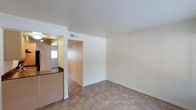 This single-story space features a straightforward, functional layout with clean lines and neutral finishes throughout. The main living area is open and unfurnished, with light beige walls and uniform tile flooring that extends across the space, providing a cohesive and easy-to-maintain surface. The ceiling is smooth and white, contributing to a bright, airy feel, and is equipped with a visible HVAC vent for climate control.

A structural dividing wall separates the living area from an adjacent hallway, which leads further into the unit. To the left, a partial view of the kitchen reveals granite or stone-look countertops and overhead lighting, suggesting a practical cooking space. A window in the kitchen area allows natural light to filter through, brightening the adjoining rooms.

The hallway, finished in warm tan tones, connects the main living space to additional areas within the unit. Overall, the space presents a clean, move-in-ready environment with a neutral palette that offers flexibility for a variety of furnishing and décor styles.