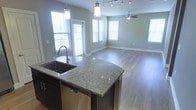 This single-floor space is thoughtfully designed with an open-concept layout that promotes a sense of flow and connectivity between living areas. The kitchen serves as the centerpiece, anchored by a curved granite island countertop with rich green and gold tones, complemented by dark cabinetry and a stainless steel undermount sink with a modern pull-down faucet. A stainless steel dishwasher is seamlessly integrated into the island, adding to the kitchen's functionality. Overhead, pendant lighting with cylindrical shades provides focused illumination above the island, while a track lighting system along the ceiling extends into the adjoining living and dining area. The living space is generous and open, featuring warm hardwood flooring that runs throughout and soft, neutral-toned walls that create a calm and welcoming ambiance. A ceiling fan is centrally positioned in the living area, adding both comfort and style. The space benefits from abundant natural light, with multiple windows dressed in white horizontal blinds, including a glass door that opens to what appears to be a balcony or exterior area. The combination of modern finishes, natural light, and open layout makes this a highly functional and inviting space.