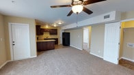 This single-floor unit showcases an open-concept design that seamlessly connects the living area and kitchen, creating a spacious, functional layout. The living area is finished with wall-to-wall carpet in a neutral tone, providing a comfortable and cohesive foundation throughout the space. A ceiling fan with a built-in light fixture is centrally positioned overhead, offering both airflow and ambient lighting to the room.

The kitchen is outfitted with rich, dark wood cabinetry that provides ample storage, complemented by a matching breakfast bar that serves as a natural dividing element between the kitchen and living areas. Black appliances, including a full-size refrigerator, add a consistent, modern aesthetic. A sink is integrated into the countertop along the back wall, and recessed-style flush-mount lighting illuminates the kitchen zone effectively.

The walls throughout are painted in a soft, warm neutral tone, contributing to an inviting and clean atmosphere. A doorway visible from the main living space leads to an additional room, suggesting further functional square footage beyond the open living and kitchen areas. Overall, the space presents a practical and well-organized interior suitable for everyday living.