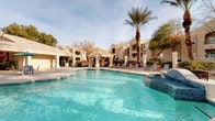 This inviting outdoor amenity area showcases a resort-style design centered around a generously sized swimming pool with crystal-clear turquoise water. A tiled in-pool fountain feature adds visual interest and a sense of tranquility to the main pool area. Adjacent to the pool, a dedicated spa offers a bubbling hydrotherapy experience, finished with decorative tile work and equipped with entry handrails for easy access.

The surrounding pool deck provides ample space for lounging, with multiple chaise lounge chairs and shaded seating areas positioned throughout. A covered pavilion with columned architecture offers a comfortable retreat from the sun, furnished with round tables and chairs ideal for outdoor dining or socializing. The Mediterranean-inspired architecture of the surrounding buildings, characterized by stucco exteriors, arched details, and tile rooflines, creates a cohesive and elegant aesthetic.

Mature palm trees and desert-adapted landscaping frame the entire space, adding both shade and a lush, tropical ambiance. A secured iron gate with decorative detailing controls access to the pool area, ensuring privacy and safety. In the background, dramatic mountain views rise above the rooftops, providing a stunning natural backdrop that enhances the overall outdoor experience.