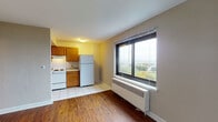 This single-floor unit offers a practical and well-organized layout spread across a living area, kitchen, two bedrooms, and a bathroom. The living area is finished with rich hardwood flooring that extends throughout most of the unit, creating a warm and cohesive feel. A large picture window with a radiator beneath it allows natural light to pour in while framing scenic views of the surrounding landscape and greenery below.

The kitchen is located adjacent to the living area and is finished with white tile flooring, warm oak cabinetry, and ample upper and lower storage. It is equipped with a white electric coil-top range, a built-in range hood, a double stainless steel sink with a standard faucet, and a full-size refrigerator. The countertops provide functional workspace, and the layout makes efficient use of the available area.

Both bedrooms feature the same hardwood flooring as the living area, with windows offering natural light and outdoor views. One bedroom includes a ceiling fan with a light fixture, adding to its comfort. Radiator heating units are present in the main rooms, providing consistent warmth throughout.

The bathroom is clean and straightforward, with white tile flooring, a toilet, a vanity with a sink, and a towel bar and toilet paper holder fitted in brushed nickel finishes. Overall, the unit presents a functional and livable space with consistent finishes throughout.