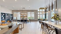 Located at 701 Watkins View Dr in Midlothian, VA, this single-floor communal space offers a thoughtfully designed open-plan layout that combines a fully equipped kitchen, dining area, and lounge into one cohesive environment. Terrazzo tile flooring runs throughout, providing a clean, contemporary foundation that complements the overall modern aesthetic.

The kitchen is outfitted with crisp white shaker cabinetry, a bold geometric hexagonal tile backsplash, stainless steel appliances including a full-size refrigerator, and a spacious island with counter seating. Large glass doors connect the kitchen directly to the outdoors, flooding the space with natural light.

The dining area features a substantial wood-top table surrounded by sleek black chairs, anchored beneath an industrial-style pendant light fixture. Built-in slate-blue bookshelves line one wall, adding both function and character, while a large abstract art panel provides a striking visual focal point.

A sun-drenched lounge nook enclosed by floor-to-ceiling windows showcases a dramatic sputnik chandelier and a round table with upholstered chairs—ideal for casual gatherings. Throughout the space, soaring ceilings with recessed lighting and multiple curated seating zones create an inviting, versatile atmosphere suited for socializing, dining, or relaxing.