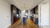 This single-floor residence showcases a thoughtfully designed interior with high-quality finishes throughout. The kitchen serves as the centerpiece of the home, featuring rich dark wood cabinetry extending floor to ceiling, granite countertops, and a full suite of stainless steel appliances including a refrigerator, range, built-in microwave, and dishwasher. A center island provides additional prep space and casual seating, while a subtle tile backsplash and recessed track lighting add refined detail. Warm hardwood flooring flows throughout the kitchen and adjoining areas, complementing the dark cabinetry beautifully. The kitchen opens toward a dining area illuminated by elegant pendant lighting, with French doors allowing natural light to fill the space. The bedroom offers a comfortable, carpeted retreat with multiple windows fitted with blinds, providing ample natural light and a bright, airy ambiance. The bathroom is finished with clean, large-format tile flooring, dark wood vanity cabinetry, granite countertops, a framed mirror, and a tub-shower combination with striking marble-style tile surround. The overall design balances contemporary style with functional living, creating a cohesive and welcoming environment throughout.
