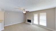 This single-story Fort Worth property offers a comfortable and functional layout highlighted by a spacious living area. The room features neutral beige carpeting that extends throughout, complementing the warm, light gray walls and crisp white baseboards. A textured popcorn ceiling adds a classic touch overhead, while a ceiling fan with integrated lighting provides both air circulation and ambient illumination.

The fireplace serves as a natural focal point, featuring a light-toned brick surround with a raised hearth and a clean white mantel that adds a traditional aesthetic to the space. Adjacent to the fireplace, built-in open shelving offers convenient display or storage options, adding architectural character to the room.

Natural light flows generously into the living area through a pair of large windows fitted with white horizontal blinds, alongside a full-length glass door that provides direct access to the outdoors. This combination of natural light and neutral finishes creates a bright, open atmosphere. Overall, the space presents a well-balanced blend of functional features and understated design, making it a welcoming environment in Fort Worth.