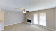 This single-story Fort Worth property offers a comfortable and functional layout highlighted by a spacious living area. The room features neutral beige carpeting that extends throughout, complementing the warm, light gray walls and crisp white baseboards. A textured popcorn ceiling adds a classic touch overhead, while a ceiling fan with integrated lighting provides both air circulation and ambient illumination.

The fireplace serves as a natural focal point, featuring a light-toned brick surround with a raised hearth and a clean white mantel that adds a traditional aesthetic to the space. Adjacent to the fireplace, built-in open shelving offers convenient display or storage options, adding architectural character to the room.

Natural light flows generously into the living area through a pair of large windows fitted with white horizontal blinds, alongside a full-length glass door that provides direct access to the outdoors. This combination of natural light and neutral finishes creates a bright, open atmosphere. Overall, the space presents a well-balanced blend of functional features and understated design, making it a welcoming environment in Fort Worth.
