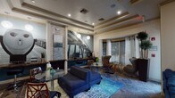 This single-floor amenity space is designed as a sophisticated communal lounge and entertainment area. The main lounge area is anchored by a large sectional sofa in deep navy blue, positioned atop a vibrant ocean-toned area rug that complements the coastal aesthetic of the space. Stone tile flooring runs throughout, adding texture and durability underfoot. A prominent stacked-stone column serves as a visual centerpiece, housing a wall-mounted flat-screen television above a dark granite ledge. A dramatic large-scale photographic mural of a pier stretches across one wall, reinforcing the coastal theme. Recessed tray ceilings with built-in lighting lend the lounge a polished, finished look. Multiple seating arrangements, including sculptural egg-style accent chairs and bistro-height tables, provide flexible options for socializing. Large windows fitted with horizontal blinds and sheer curtains allow natural light to filter through while framing outdoor views. A dedicated workstation area with computers is integrated into the lounge. An adjoining game room features a full-size billiards table beneath a statement chandelier, complemented by patterned carpeting, framed wall art, wall sconces, and additional seating, creating a well-rounded entertainment environment.