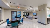 Located at 5200 S Ulster St in Greenwood Village, Colorado, this single-floor amenity space offers a well-appointed communal environment designed for both relaxation and social gatherings. The open-plan layout is anchored by large-format white tile flooring that extends throughout, creating a clean, cohesive aesthetic. Track lighting runs along the ceiling, providing flexible, well-distributed illumination across the entire space.

The lounge area serves as the centerpiece, featuring a grouping of contemporary light-grey upholstered armchairs and sofas arranged around a glass-top coffee table, all set atop a patterned area rug. A standout stacked-stone accent wall provides striking texture and serves as the backdrop for a wall-mounted flat-screen television, lending the space a modern, upscale character.

A dining zone with dark wood tables and teal-upholstered chairs offers a more structured seating option, complemented by bar-height tables and yellow-accented stools nearby. Glass doors lead directly to an outdoor patio, allowing natural light to filter in and providing easy access to the exterior.

The fully equipped galley-style kitchen features dark espresso cabinetry, white quartz countertops, stainless steel appliances, and a sink with modern fixtures. Pendant lighting and teal decorative accents tie the kitchen's design into the broader color palette of the space, making it fully functional for catered events or casual use.