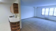This single-floor space offers an open, functional layout with a clean and straightforward design. The main living area is finished with wall-to-wall carpeting in a neutral tone, contributing to a quiet and understated ambiance. White walls throughout keep the space feeling bright and simple, while a window along one wall allows natural light to filter in.

A compact kitchenette is integrated along one side of the space, featuring a countertop with a sink, upper cabinetry with open shelving, and a lower rounded shelving unit that adds a distinctive built-in storage element. The warm wood tones of the cabinetry provide a contrast against the light walls and countertop surfaces.

A structural column is visible near the far end of the main area, and a baseboard heating unit runs along the base of the exterior wall beneath the window. The overall layout is open and unobstructed, offering flexibility in how the space can be arranged or utilized. The design is practical and modest, suited for straightforward, everyday use.