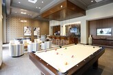 This single-floor amenity space is thoughtfully designed to accommodate a variety of social and professional activities, featuring distinct zones that flow seamlessly together. The game room anchors one end of the space, showcasing a full-size pool table set against floor-to-ceiling windows that flood the area with natural light and offer views of the surrounding streetscape. Tall, draped curtains frame the glazed walls, adding warmth to the otherwise modern aesthetic, while a wall-mounted television and geometric pendant lighting complete the room.

Adjacent to the game room, a media lounge offers an intimate retreat with high-backed upholstered chairs arranged around a flat-screen television set into a rich walnut wood accent wall. A bold patterned wallcovering and large abstract artwork contribute texture and visual interest to the space.

A dedicated conference area features a triangular wood table surrounded by contemporary chairs, patterned wallcovering, and access to natural light through floor-to-ceiling windows. A centrally located wet bar with stone countertops and bar seating serves as the social hub connecting the zones. Throughout the space, carpeted flooring, elevated ceilings, and cohesive warm-toned finishes create a polished, welcoming ambiance.