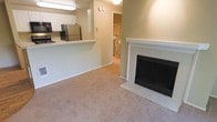 This single-floor residence offers a functional and comfortable layout designed for easy everyday living. The living area is carpeted in neutral tones, creating a warm and inviting atmosphere, and is anchored by a classic fireplace featuring a white tile surround and mantel — a standout focal point of the space. The open-concept design seamlessly connects the living area to the kitchen, which is accessible via a raised breakfast bar or pass-through counter. The kitchen is equipped with stainless steel appliances, including a refrigerator and microwave, and features white cabinetry that provides ample storage. The walls throughout are painted in soft, neutral green-beige tones, contributing to a calm and cohesive aesthetic. A hallway visible from the main living space leads to additional areas, including what appears to be a utility or laundry area with a water heater, suggesting practical storage solutions within the home. The overall layout is efficient and well-organized, offering a blend of comfort and functionality suited to a variety of lifestyles.