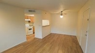 This single-story space features an open-concept layout that connects the living area to the kitchen, creating a functional and cohesive floor plan. The living area is finished with warm hardwood-style flooring that extends throughout, adding continuity and a welcoming feel to the space. A ceiling fan with a built-in light fixture is centrally mounted, providing both air circulation and ambient lighting.

A partial wall with a pass-through counter separates the living area from the kitchen, maintaining an open feel while defining each zone. The kitchen is equipped with warm-toned wood cabinetry, a white electric range, and an overhead ventilation hood. A HVAC vent is visible near the kitchen entrance, indicating a central air system. The cabinetry offers ample storage, and the layout allows for efficient use of the cooking space.

The walls are finished in a neutral off-white tone throughout, complementing the flooring and cabinetry. The textured ceiling adds a subtle design detail. Overall, the space offers a straightforward, functional design with consistent finishes that create a cohesive and comfortable environment.