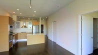 This single-floor space showcases a clean, contemporary design that emphasizes open living and functional elegance. The open-concept layout seamlessly connects the kitchen and living area, creating a sense of spaciousness and flow. The kitchen is well-appointed with modern flat-front cabinetry in a warm wood tone, complemented by light upper cabinets and a generous island with a light countertop that serves as both a workspace and casual gathering spot. A stainless steel refrigerator and professional-style range hood anchor the culinary space, lending it a polished, efficient character. Recessed lighting and track lighting are thoughtfully placed throughout the ceiling, providing ample illumination while maintaining a streamlined aesthetic. The rich, dark hardwood floors run continuously across the space, adding warmth and contrast to the bright white walls. The living area is generous and versatile, framed by clean, smooth walls that offer a blank canvas for personalization. A minimalist interior door with a flush design reinforces the modern sensibility found throughout. Overall, the space balances contemporary style with practical design, making it both visually appealing and highly livable.