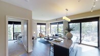 This single-floor residence showcases a thoughtfully designed open-concept layout that seamlessly connects the kitchen, dining, and living areas into one cohesive living space. The kitchen features a substantial granite-topped island with bar seating, dark cabinetry, and a stylish rectangular pendant light fixture that anchors the space. The island serves as both a functional workspace and a natural divider between the kitchen and the adjoining dining and living areas.

The living room is generously sized, furnished with a neutral sectional sofa, an accent armchair, and a coffee table set atop a patterned area rug over rich dark hardwood flooring. Large floor-to-ceiling windows along two walls flood the space with abundant natural light while providing open views of the surrounding tree-lined neighborhood. The soft beige and warm grey tones of the walls complement the contemporary furnishings throughout.

A frosted glass door near the entry adds a modern architectural detail while maintaining privacy to adjoining rooms. The overall ambiance is clean, bright, and contemporary, combining functional design with an inviting, comfortable atmosphere.