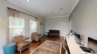 Located at 926 Cleveland St in Greenville, SC, this single-room office offers a polished and functional workspace with a warm, professional ambiance. The space is defined by its rich, dark hardwood floors that run throughout, complemented by neutral gray walls, crisp white crown molding, and clean white baseboards that give the interior a refined, cohesive look.

Natural light pours in through multiple windows dressed with horizontal blinds and sheer linen-toned curtains, creating a bright and inviting atmosphere. Recessed lighting in the ceiling provides consistent overhead illumination throughout the day.

The layout is thoughtfully arranged to support both productivity and visitor comfort. A long, built-in white desk with dark wood cabinetry below provides an organized workstation area, while a compact lounge seating arrangement — featuring upholstered chairs, small side tables, and an accent stool — creates a welcoming area for meetings or consultations. Framed abstract artwork adorns the walls, adding a touch of creativity to the professional setting.

A glass-paneled interior door connects to an adjoining space, enhancing the sense of openness. The overall design balances practicality with comfort, making this office in downtown Greenville well-suited for a variety of professional uses.