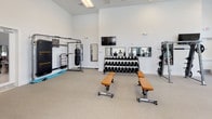 This single-level fitness center is designed to support a full spectrum of workout needs within a bright, open environment. The space is characterized by soaring double-height ceilings that create an airy, expansive atmosphere, complemented by large floor-to-ceiling windows and a row of circular porthole-style windows that flood the interior with natural light. Roller shades provide adjustable light control throughout the day, while a ceiling fan aids in air circulation.

The flooring consists of durable, speckled rubberized material suited for heavy equipment use. The cardio zone is well-appointed with Matrix-brand elliptical trainers, treadmills, and a rowing machine, all positioned to take advantage of the outdoor views. A dedicated strength training area includes a multi-station cable machine system, a full rack of dumbbells ranging across multiple weight increments, adjustable benches, a lat pulldown station, and a selection of selectorized resistance machines with tan upholstered seating.

A functional training corner features a punch bag, wall-mounted agility tools, and a flat-screen television mounted on the crisp white wall. Wall mirrors are positioned strategically throughout to support form checking. The overall aesthetic is clean and modern, maintaining a professional gym atmosphere.