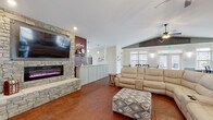 This single-floor gathering space is designed for comfort and functionality, offering an open and inviting layout throughout. The main living area is anchored by a striking floor-to-ceiling stacked stone fireplace with an electric insert and a large wall-mounted TV, set against a rich, deep red accent wall. Plush reclining sofas and an ottoman create a relaxed seating arrangement, all resting on richly toned stamped concrete flooring that flows seamlessly throughout the space. Vaulted ceilings with dual ceiling fans add to the airy, expansive feel of the room. The dining area accommodates large groups with multiple dark wood farmhouse-style tables and cross-back chairs, bathed in natural light from generously sized windows trimmed with classic white wainscoting and two-tone painted walls. The fully equipped kitchen features light gray shaker cabinetry, granite countertops, a center island with bar seating, stainless steel appliances including a range with a sleek hood vent, double wall oven, and dishwasher, all set against a bold red accent wall. A set of glass-panel double doors leads to an outdoor pool area, visible through large interior windows, adding a bright and recreational dimension to this well-appointed communal space.