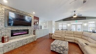 This single-floor gathering space is designed for comfort and functionality, offering an open and inviting layout throughout. The main living area is anchored by a striking floor-to-ceiling stacked stone fireplace with an electric insert and a large wall-mounted TV, set against a rich, deep red accent wall. Plush reclining sofas and an ottoman create a relaxed seating arrangement, all resting on richly toned stamped concrete flooring that flows seamlessly throughout the space. Vaulted ceilings with dual ceiling fans add to the airy, expansive feel of the room. The dining area accommodates large groups with multiple dark wood farmhouse-style tables and cross-back chairs, bathed in natural light from generously sized windows trimmed with classic white wainscoting and two-tone painted walls. The fully equipped kitchen features light gray shaker cabinetry, granite countertops, a center island with bar seating, stainless steel appliances including a range with a sleek hood vent, double wall oven, and dishwasher, all set against a bold red accent wall. A set of glass-panel double doors leads to an outdoor pool area, visible through large interior windows, adding a bright and recreational dimension to this well-appointed communal space.