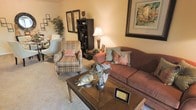 This single-floor space features a welcoming living room designed with comfort and classic style in mind. The living area is furnished with a rich, earthy-toned sofa accented by decorative throw pillows in soft pink and sage green hues, complemented by a striped upholstered armchair with a coordinating ottoman. A traditional wooden coffee table serves as a central focal point, adorned with decorative figurines and potted greenery. Warm ambient lighting is provided by a classic table lamp, casting a soft glow throughout the room. The walls are dressed with framed artwork, adding visual interest and a cultured ambiance to the space. A dark wood bookcase provides both storage and display space, while lush indoor plants introduce a natural, organic element. Plush wall-to-wall carpeting in a neutral tone runs throughout, enhancing the overall sense of warmth and coziness. The space has a well-curated, lived-in feel with thoughtful decorative touches that create an inviting and comfortable atmosphere suitable for relaxing or entertaining.