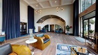 This single-floor community clubhouse and leasing lounge is defined by its striking architectural presence and thoughtfully curated interior design. The grand entry lobby immediately draws attention with its soaring coffered ceiling, dramatic arched architectural detail, and floor-length navy curtains that frame the space with elegance. Warm hardwood flooring flows throughout, complementing the neutral wall tones and contemporary decor. Statement pendant light fixtures with exposed Edison-style bulbs hang from the ceiling, adding an artistic, modern-industrial touch.

The reception and leasing area is visible through the arched opening, featuring a dedicated desk area with seating for staff, surrounded by lush potted greenery. Sculptural accent pieces, including a notable horse bust atop a slender side table, contribute to the space's distinctive character.

Adjacent lounge areas offer generous seating arrangements, including a plush navy velvet sofa positioned beside large floor-to-ceiling windows that invite abundant natural light and frame views of the surrounding landscaped grounds. A second seating area features a channeled sectional sofa, glass coffee table, and emerald green accent chair, all set beneath towering windows with sheer drapery. A wet bar with wood cabinetry and gold hardware adds a refined, functional element to the lounge.