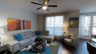 This single-floor space centers around a thoughtfully designed living room that balances comfort and modern aesthetics. The room features a large sectional sofa in a neutral gray tone, complemented by blue accent pillows that add a pop of color. A matching chaise lounge extends the seating arrangement, creating an inviting area for relaxation.

At the heart of the room sits a round glass-top coffee table with a dark base, adorned with decorative items including a sculptural rhinoceros figurine and reading material. A teal and white area rug anchors the seating arrangement atop rich, dark hardwood flooring, adding texture and visual interest to the space.

Natural light filters generously through a large window dressed with sheer white curtains and horizontal blinds, creating a bright and airy atmosphere. A ceiling fan with a built-in light fixture is centrally positioned, enhancing both air circulation and ambient lighting. Wall art in warm and cool tones is thoughtfully displayed, contributing to the room's curated feel. A floor lamp beside a sleek accent chair in the corner provides additional warm lighting, completing this well-balanced living space.