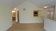 This single-story space centers around an open living area distinguished by a vaulted, angled ceiling that adds a sense of height and volume to the room. The space is finished with wall-to-wall carpet in a neutral beige tone, contributing to a warm and understated aesthetic throughout. Cream-colored walls keep the interior feeling bright and clean.

A built-in pass-through window connects the living area to the kitchen, offering a practical open-concept feel without fully removing the wall between the two spaces. Through the pass-through, the kitchen is partially visible, revealing wood-toned cabinetry, a black microwave, and what appears to be a range — providing a functional cooking space with standard appliances. A ventilation register is positioned high on the shared wall between the living area and kitchen, indicating a central HVAC system.

To the left side of the living area, a hallway leads to additional rooms, with a bathroom visible at the end featuring a vanity with wood cabinetry and a wall-mounted light fixture. The overall layout is straightforward and efficient, with a cohesive, neutral design palette throughout.