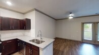 This single-story residence in Magnolia, Texas offers a thoughtfully designed layout with modern finishes throughout. The open-concept kitchen is a standout feature, boasting rich dark-stained cabinetry, granite countertops with a double stainless steel sink, and a full suite of stainless steel appliances including a gas range, microwave, and French door refrigerator with ice maker. Recessed lighting illuminates the kitchen, which flows seamlessly into the living area via a peninsula-style counter. The living and dining area features warm wood-look flooring, crown molding, and a ceiling fan, creating a comfortable and cohesive space. The bedroom is carpeted for added comfort, filled with natural light from a large window with views of lush greenery, and includes a ceiling fan. The bathroom is clean and functional, appointed with a glass-enclosed walk-in shower with tile surround, wood-look flooring, and chrome grab bars and fixtures. Throughout the unit, neutral tones, crown molding, and consistent flooring choices contribute to a polished, move-in-ready aesthetic suited for comfortable everyday living.