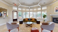 Located in Austin, TX, this single-story clubhouse offers a thoughtfully designed gathering space that blends architectural elegance with functional amenity areas. The centerpiece of the clubhouse is a stunning circular lounge defined by a sweeping wall of floor-to-ceiling windows that flood the space with natural light and frame picturesque views of the outdoor pool and mature surrounding trees. A coffered, octagonal tray ceiling adds architectural depth overhead, while warm beige tile flooring flows throughout. A classic white-mantled fireplace anchors one end of the living area, complemented by curated seating including a plush velvet sofa and accent chairs arranged for both comfort and conversation. Adjacent to the lounge, a formal dining area benefits from soaring arched windows dressed with floor-length drapes, structural columns with clean white millwork, and a welcoming entry door. The kitchen is outfitted with rich dark wood cabinetry, granite countertops, a mosaic tile backsplash, and stainless steel appliances, all positioned to overlook the pool area through an arched pass-through window. A dedicated office space with hardwood-style flooring and a built-in desk area rounds out the clubhouse, offering a functional workspace within the broader community facility.