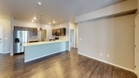 This single-floor residence presents a cohesive, modern design throughout its various living spaces. The open-concept kitchen and living area serves as the heart of the home, where rich dark wood cabinetry pairs with light quartz-style countertops and a full suite of stainless steel appliances, including a side-by-side refrigerator with ice maker, electric range, and over-the-range microwave. Warm-toned pendant lighting illuminates the kitchen peninsula, which overlooks the spacious living area. Dark hardwood-style flooring runs throughout the main living spaces, complementing the neutral warm-gray wall tones. The living room benefits from natural light via French doors that open to an exterior space. The entry foyer provides a clean transition into the home with additional storage nearby. The bedrooms feature plush carpeting and windows that bring in natural light, offering comfortable, quiet retreats. Two bathrooms serve the unit: a full bath with a tub/shower combination, dark vanity cabinetry, stone-look countertops, and wood-style flooring; and a second bath with tile-style flooring, matching dark cabinetry, and additional upper cabinet storage. Overall, the space maintains a consistent, contemporary aesthetic with practical finishes throughout.