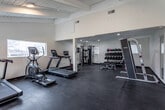 Located at 5408 E 96th Place in Kansas City, MO, this single-floor fitness facility offers a comprehensive workout environment designed to support a wide range of exercise routines. The space is thoughtfully laid out across an open floor plan, with dark rubber flooring throughout that provides both durability and traction underfoot.

The facility is equipped with multiple cardio machines, including Star Trac treadmills, a spin bike with a wall-mounted display screen, and a rowing machine. Strength training options include a multi-function cable machine, a Smith machine with an adjustable bench, and a well-stocked dumbbell rack featuring a variety of weights and medicine balls. The varied equipment arrangement allows for efficient use of the space without feeling crowded.

Architectural features include exposed beam ceilings in portions of the facility, recessed lighting, and large windows along the exterior wall that flood the space with natural light and offer views of the surrounding outdoor area, which appears to include a pool and open landscape. A glass-paneled entry door provides access to the outdoor space. The clean, neutral-toned walls and open layout contribute to a functional, motivating atmosphere well-suited for personal training or general fitness use.
