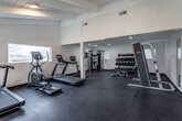 Located at 5408 E 96th Place in Kansas City, MO, this single-floor fitness facility offers a comprehensive workout environment designed to support a wide range of exercise routines. The space is thoughtfully laid out across an open floor plan, with dark rubber flooring throughout that provides both durability and traction underfoot.

The facility is equipped with multiple cardio machines, including Star Trac treadmills, a spin bike with a wall-mounted display screen, and a rowing machine. Strength training options include a multi-function cable machine, a Smith machine with an adjustable bench, and a well-stocked dumbbell rack featuring a variety of weights and medicine balls. The varied equipment arrangement allows for efficient use of the space without feeling crowded.

Architectural features include exposed beam ceilings in portions of the facility, recessed lighting, and large windows along the exterior wall that flood the space with natural light and offer views of the surrounding outdoor area, which appears to include a pool and open landscape. A glass-paneled entry door provides access to the outdoor space. The clean, neutral-toned walls and open layout contribute to a functional, motivating atmosphere well-suited for personal training or general fitness use.