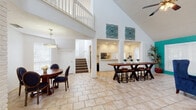 This two-story space makes an immediate impression with its dramatic vaulted ceilings that rise through both floors, creating a sense of openness throughout the main level. Light-toned tile flooring flows across the ground floor, connecting a series of well-defined living and dining areas. The focal point of the main living area is a bold teal-painted brick fireplace wall, topped with a wall-mounted television and flanked by plush navy blue wingback chairs — a striking combination of color and texture that anchors the room. Ceiling fans are positioned throughout, complementing the airy atmosphere.

Adjacent to the lounge area, a large communal dining table with bar-height seating provides ample space for group gatherings, while a separate round dining table with upholstered chairs sits beside a white-painted brick accent wall. A full-size pool table occupies its own dedicated zone near French doors that invite natural light from outside.

The galley-style kitchen is efficiently laid out with white cabinetry, light quartz countertops, and stainless steel appliances including a range and microwave.

A staircase with white wooden railings leads to the second floor, where a carpeted loft and open hallway overlook the main level below. The upper floor also features vaulted ceilings, a dining area with wicker seating, and additional flex space, making it a versatile extension of the overall layout.