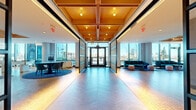 Located at 180 Morgan Street in Jersey City, NJ, this sophisticated amenity space spans a generously sized single floor, offering a diverse range of social and recreational areas designed for modern living. The entrance corridor immediately impresses with its striking herringbone-patterned flooring, a warm coffered wood ceiling with recessed and pendant lighting, and floor-to-ceiling glass walls that flood the space with natural light and frame panoramic views of the surrounding cityscape.

The open-plan layout flows seamlessly from a welcoming lobby into distinct functional zones. A lounge area features teal velvet sofas, pink ottomans, and a bar counter with brass pipe detailing and high stools, all bathed in natural light from expansive windows. Adjacent to this is a co-working and dining area with long communal tables, upholstered navy chairs, and arched mirror-panel wall accents that add depth and elegance.

The game room is a standout feature, housing a sleek white-and-gold shuffleboard table alongside a billiards table, set against a backdrop of sweeping floor-to-ceiling windows overlooking the Jersey City skyline. Throughout the space, the consistent use of cork-tile flooring, bold blue area rugs, and warm wood ceiling elements creates a cohesive, upscale aesthetic that balances comfort with contemporary design.