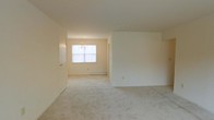 This single-floor residence offers a clean, understated aesthetic defined by its warm neutral palette and soft, plush wall-to-wall carpeting. The open living and dining areas flow seamlessly into one another, creating a generous and versatile layout well-suited for both everyday living and entertaining. Natural light filters through a central window in the dining area, brightening the space and lending an airy, welcoming atmosphere to the interior. An elegant chandelier serves as a focal point overhead, adding a refined touch to the dining area. Baseboard heating runs along the perimeter, ensuring comfort throughout the seasons. The smooth white walls and ceilings provide a blank canvas adaptable to a variety of décor styles. A doorway connects the main living space to an adjacent room, offering a practical and intuitive flow between areas. The overall ambiance is calm and understated, with a functional, move-in-ready quality that makes the space immediately livable. The consistent use of light tones across the floors, walls, and ceilings contributes to a cohesive and harmonious environment throughout the home.