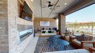 This covered outdoor amenity space offers a well-appointed gathering area designed for both relaxation and socializing. The architecture is defined by warm-toned exposed brick walls and sturdy brick columns that frame open-air views of the surrounding landscaped grounds and community buildings beyond. A paneled ceiling overhead is equipped with ceiling fans and recessed lighting, ensuring comfort throughout the day and into the evening.

The lounge area is generously furnished with a mix of plush sofas, wood-framed armchairs with neutral cushions, and accent pillows in deep greens, all arranged over a bold patterned area rug. Sleek round side tables provide functional surfaces throughout the seating zone. A linear fireplace is built into one wall alongside a large wall-mounted television, creating a focal point ideal for casual gatherings. Wall art adds a decorative touch against the brick backdrop.

A separate dining zone features a long natural wood communal table surrounded by black mesh chairs, perfect for group meals or collaborative use. An additional bistro table with chairs extends onto the outer edge of the patio. The space opens broadly on multiple sides, allowing abundant natural light and unobstructed sightlines to the outdoors, making it a versatile and inviting retreat for residents.