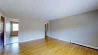 This single-floor space offers a straightforward and well-maintained layout designed for comfortable everyday living. The main living area is immediately welcoming, showcasing warm honey-toned hardwood floors that run throughout, adding a sense of continuity and warmth to the space. The walls are finished in a soft, neutral gray-white tone that reflects light evenly and provides a versatile backdrop for a variety of interior styles. White baseboards frame the perimeter of the room cleanly, complementing the overall minimal aesthetic. A doorway visible from the main room leads into an adjoining area, suggesting a connected and practical floor plan that allows for easy movement throughout the space. Baseboard heating units are positioned along the walls, providing an efficient and unobtrusive heating solution. The smooth, white ceiling contributes to an open and airy feel. The space is clean, neutral, and move-in ready, making it well-suited for those who value simplicity and functionality. Overall, the design prioritizes clean lines and a calm, understated atmosphere throughout.