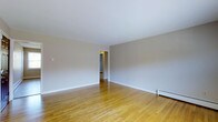 This single-floor space offers a straightforward and well-maintained layout designed for comfortable everyday living. The main living area is immediately welcoming, showcasing warm honey-toned hardwood floors that run throughout, adding a sense of continuity and warmth to the space. The walls are finished in a soft, neutral gray-white tone that reflects light evenly and provides a versatile backdrop for a variety of interior styles. White baseboards frame the perimeter of the room cleanly, complementing the overall minimal aesthetic. A doorway visible from the main room leads into an adjoining area, suggesting a connected and practical floor plan that allows for easy movement throughout the space. Baseboard heating units are positioned along the walls, providing an efficient and unobtrusive heating solution. The smooth, white ceiling contributes to an open and airy feel. The space is clean, neutral, and move-in ready, making it well-suited for those who value simplicity and functionality. Overall, the design prioritizes clean lines and a calm, understated atmosphere throughout.