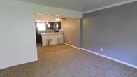 This single-floor residence offers a functional and comfortable layout designed for everyday living. The open-concept living area is finished with plush carpet flooring and painted in cool grey tones, creating a calm and neutral ambiance throughout. Crown molding adds a subtle decorative touch along the ceiling line, lending a polished finish to the space.

The kitchen is positioned adjacent to the living area and is defined by a raised breakfast bar countertop with mosaic tile detailing, providing both a casual dining and meal prep surface. Two metal barstools are situated at the bar, making it an ideal spot for quick meals or entertaining guests. Pendant lighting hangs above the bar area, while a ceiling fan with a light fixture serves the connected living space, ensuring both illumination and air circulation. The kitchen features dark cabinetry and appears to be equipped with standard appliances, offering practical storage and workspace.

A structural beam separates the two zones while maintaining the open feel of the floor plan. White baseboards run throughout, complementing the grey walls cleanly. Natural light filters in through a window visible in the kitchen area, adding brightness to the interior. Overall, the space presents a clean, move-in ready environment with a cohesive design palette.