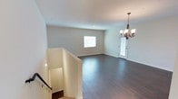 This well-appointed single-floor space showcases a thoughtful blend of modern design and functional layout. The main living area immediately draws attention with its rich, dark hardwood flooring that runs throughout, providing a striking contrast against the soft, neutral-toned walls and crisp white ceiling. The open layout creates a generous sense of space, allowing for flexible furniture arrangements and easy movement throughout the room. A classic multi-arm chandelier with warm-toned bulbs serves as an elegant focal point, adding a touch of traditional charm to the otherwise contemporary aesthetic. Natural light filters in through a centrally placed window, brightening the interior and enhancing the airy feel of the space. A glass-paneled entry door further contributes to the light-filled ambiance while providing a welcoming transition point. Clean white baseboards frame the perimeter of the room, adding a polished finishing detail. The staircase landing visible in the imagery features a sleek railing and a subtle recessed light, offering a smooth architectural transition. Overall, the space presents a pristine, move-in-ready environment with a neutral palette that lends itself well to a variety of interior design styles.