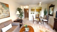 This single-floor residence offers a warm and functional layout that connects the living, dining, and kitchen areas in a cohesive, comfortable flow. The living room is a welcoming space featuring light neutral carpet, a ceiling fan, and room for generous seating arrangements. Lush greenery and soft lamp lighting contribute to an inviting, homey ambiance.

The dining area is thoughtfully positioned between the living room and kitchen, offering flexibility with space for both a formal dining table and a cozy bistro-style secondary seating nook. A pass-through window connects the dining area directly to the kitchen, enhancing the open feel and making the space ideal for entertaining.

The kitchen is compact yet well-appointed, featuring warm wood-toned upper and lower cabinetry, light countertops, a stainless steel sink, and full appliances including a range and refrigerator. Tile flooring in the kitchen provides a practical transition from the carpeted living spaces.

A entry hallway with a wood-paneled door provides a defined arrival point, adding structure to the overall layout. Throughout the residence, neutral wall tones and soft carpeting create a calm, understated aesthetic. The design balances everyday practicality with a comfortable, lived-in character that makes the space feel both functional and welcoming.