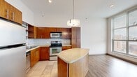 This single-floor residence offers a thoughtfully designed layout encompassing one bedroom, two bathrooms, a combined kitchen and living area, a dedicated laundry room, and a closet. The open-concept kitchen and living space serves as the heart of the home, featuring warm wood cabinetry, a subway tile backsplash, granite countertops, and a full suite of stainless steel appliances including a range, microwave, and refrigerator. A curved kitchen island with a matching granite surface provides additional workspace and casual seating. The flooring transitions from tile in the kitchen to hardwood-style planks in the living area, creating visual definition between the two zones. The living area is bathed in natural light through large, floor-to-ceiling windows fitted with adjustable blinds, offering open city views. The bedroom features plush carpeting and a window that draws in additional natural light. Both bathrooms are finished with wood vanities, granite countertops, and modern light fixtures. The dedicated laundry room includes a full-size washer and dryer with overhead shelving. Throughout the residence, high ceilings, clean neutral walls, and consistent modern finishes contribute to an open, airy, and functional living environment.