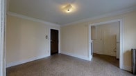 This single-floor space presents a well-maintained interior with a timeless aesthetic. The main living area is finished with speckled terrazzo flooring, which adds both durability and a classic visual texture underfoot. Walls are painted in a warm, neutral cream tone and are accented by crisp white crown molding that runs along the ceiling perimeter, lending an air of traditional elegance to the space.

A dark wood entry door serves as a focal point along one wall, providing a strong contrast against the light-colored surroundings. Overhead, a flush-mount ceiling light fixture provides ambient illumination throughout the room. An arched doorway transitions into an adjoining area, adding an architectural detail that softens the overall layout and contributes to the space's classic character. A partial wall divider with a white-painted finish separates portions of the space while maintaining an open feel.

The adjoining area, visible through the arch, features additional cabinetry and a door, suggesting storage or utility functionality. The overall ambiance is clean, understated, and move-in ready, with architectural details that reflect a well-preserved traditional design.