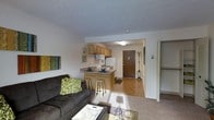 This single-floor unit offers an efficient and functional open-concept layout that seamlessly connects the living area to the kitchen. The living space is carpeted and accommodates a comfortable seating arrangement, complemented by natural light that brightens the interior. A framed wall features colorful artwork, adding a touch of personality to the space.

The kitchen is positioned at the heart of the unit, equipped with light wood cabinetry that provides generous storage, a white refrigerator, a gas range, and a sink positioned beneath upper cabinets. A peninsula with a dark countertop extends into the space, creating a breakfast bar with seating for two bar stools — ideal for casual dining or additional workspace.

The entryway features hard flooring and is accessed through a solid wood front door, with a coat rack nearby for added convenience. Adjacent to the entry, a well-organized open closet offers practical storage solutions. The space maintains a clean, neutral color palette throughout, with white walls and a textured ceiling that contribute to a bright and airy ambiance. Overall, the layout is designed to maximize functionality within a cozy, single-level footprint.