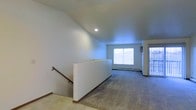 This single-floor space presents a clean, open layout designed for comfortable everyday living. The main living area is finished with wall-to-wall carpeting that adds warmth underfoot, while crisp white walls and recessed ceiling lighting create a bright, airy atmosphere throughout.

A prominent staircase with a wooden handrail is positioned toward one end of the space, featuring a low half-wall enclosure that defines the stairwell without closing off the room. Natural light flows generously into the space through a large window at the far end, enhancing the sense of openness and depth.

A baseboard heating unit runs along the exterior wall beneath the window, providing an efficient and unobtrusive climate control solution. An additional entry point with a glass-panel door on the right side of the space offers further access and allows additional natural light to filter in.

The overall ambiance is understated and neutral, with a simple, functional design that provides a versatile foundation. The combination of natural light, open sightlines, and straightforward finishes makes this a practical and adaptable space suited to a variety of uses.