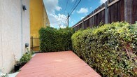 This single-story property includes a notable outdoor deck area that serves as a defining feature of the space. The deck is constructed with warm-toned wooden planks, offering a clean and functional surface well-suited for outdoor relaxation or entertaining. Flanking both sides of the deck are dense, well-maintained hedges that form a natural green enclosure, providing a strong sense of privacy and seclusion from the surrounding urban environment. A tall, dark-stained wooden privacy fence runs along the back perimeter, further reinforcing the enclosed character of the outdoor area. On one side, a black wrought iron gate provides a secure point of entry or exit, adding a structural contrast to the organic greenery. The bright yellow exterior wall of the building is visible along one edge, contributing a bold pop of color to the setting. Above, an open sky with scattered clouds allows ample natural light to fill the space throughout the day, creating an airy atmosphere despite the enclosed perimeter. The overall ambiance is one of a private urban retreat, where lush landscaping and structured fencing combine to create a comfortable and sheltered outdoor environment.