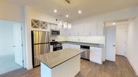 This single-floor space showcases a well-appointed, modern kitchen designed with both functionality and style in mind. The kitchen is anchored by a central island with a smooth gray countertop, providing ample workspace and serving as a natural focal point. Surrounding the island, the L-shaped layout maximizes efficiency, offering generous counter space along the perimeter.

White shaker-style cabinetry lines the walls, creating a clean, bright aesthetic that is enhanced by recessed lighting and three decorative pendant lights suspended above the island. A built-in wine rack with an X-pattern design adds a distinctive architectural detail in the upper corner, offering practical storage with visual interest.

The kitchen is fully equipped with stainless steel appliances, including a refrigerator, gas range, microwave, and dishwasher, all seamlessly integrated into the cabinetry. A stainless steel sink with a modern faucet is positioned along the perimeter counters. Wood-look flooring runs throughout, adding warmth and texture that contrasts pleasantly with the crisp white cabinetry and cool gray countertops. The overall ambiance is contemporary, clean, and highly functional.
