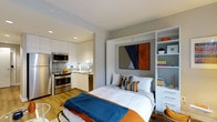 This thoughtfully designed single-floor studio maximizes every square foot through smart, space-efficient solutions. The open-plan layout connects the kitchen and living/sleeping areas in a seamless flow, creating a cohesive and functional living environment. The kitchen is outfitted with stainless steel appliances, including a full-size range, microwave, and refrigerator, complemented by sleek white cabinetry and a classic tile backsplash. Warm hardwood flooring runs throughout, adding a sense of continuity and warmth. The sleeping area features a built-in Murphy bed system framed by integrated white shelving and drawer units, offering generous storage without sacrificing style. When the bed is deployed, it serves as the room's focal point, accentuated by a bold abstract artwork in rust, navy, and grey tones mounted above the headboard. Recessed lighting throughout the ceiling provides clean, even illumination. The overall color palette — soft grey walls, white cabinetry, and pops of orange and blue in the furnishings — creates a modern, energetic, yet comfortable ambiance. This studio is a well-appointed, efficient living space ideal for those seeking contemporary urban living.