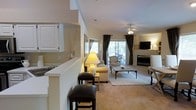 This two-story home offers a thoughtfully designed layout with a warm, inviting ambiance throughout. The main living area is an open-concept space anchored by a tile-surround fireplace, providing a natural focal point for the room. Plush carpeting, neutral wall tones, and a ceiling fan overhead contribute to a relaxed and comfortable atmosphere. A large sliding glass door floods the space with natural light and opens to an outdoor balcony, where additional seating invites enjoyment of the exterior views. The kitchen features classic white cabinetry and is separated from the living area by a raised breakfast bar, complete with bar stool seating — an ideal setup for casual dining or entertaining. The half-wall partition between the kitchen and living room maintains an open feel while subtly defining each space. Elegant drapery in deep, muted tones frames the windows and balcony door, adding a refined touch to the overall décor. The seamless flow between the kitchen, dining, and living zones makes this home well-suited for both everyday living and hosting guests. Spanning two floors, the home provides ample space and versatile living arrangements throughout.