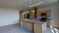 This single-floor space centers around an open-concept kitchen that seamlessly integrates with the surrounding living area. The kitchen is anchored by a generous island topped with a sleek gray countertop, offering abundant workspace along with built-in open shelving on one end, providing both functional storage and display opportunities. Warm honey-toned wood cabinetry lines the walls and base, offering a cohesive, contemporary aesthetic balanced by gray laminate countertops throughout.

Stainless steel appliances, including a refrigerator, microwave, and a gas range, are integrated into the layout, complementing the modern design. A deep sink is positioned along the island, making the space highly functional. Three industrial-style pendant lights hang above the island, providing focused task lighting, while a linear overhead fixture adds supplemental illumination. The polished concrete floors lend an urban, loft-like character to the interior. Neutral gray walls keep the palette understated, allowing the wood cabinetry to serve as the primary visual focal point. A hallway visible in the background suggests additional rooms beyond the main open area.