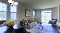 This single-floor living space offers an open, well-lit layout designed for comfortable everyday living. The living area is anchored by a neutral-toned sofa dressed with an assortment of decorative pillows in warm earth tones, set atop a bold blue patterned area rug that adds visual interest to the room. A glass-top coffee table and matching side table provide functional surfaces without overwhelming the space. Large windows along multiple walls flood the interior with natural light, while sliding glass doors at the far end open to a view of lush greenery, seamlessly connecting the indoors with the outside. The walls are finished in a soft, neutral gray tone that complements the furnishings and creates a calm, cohesive ambiance throughout. A ceiling-mounted light fixture with a classic globe design serves as a subtle focal point near the dining area, where a dark-toned table and chairs offer a defined space for meals. A framed piece of wall art adds a decorative accent, contributing to the polished, move-in-ready feel of this inviting living space.